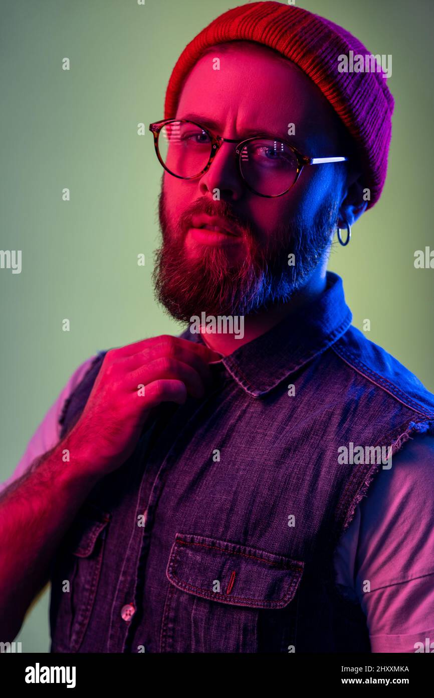 Portrait of attractive bearded hipster man in glasses, looking at camera, holding hand on his collar, wearing red beanie hat. Indoor studio shot isolated on colorful neon light background. Stock Photo