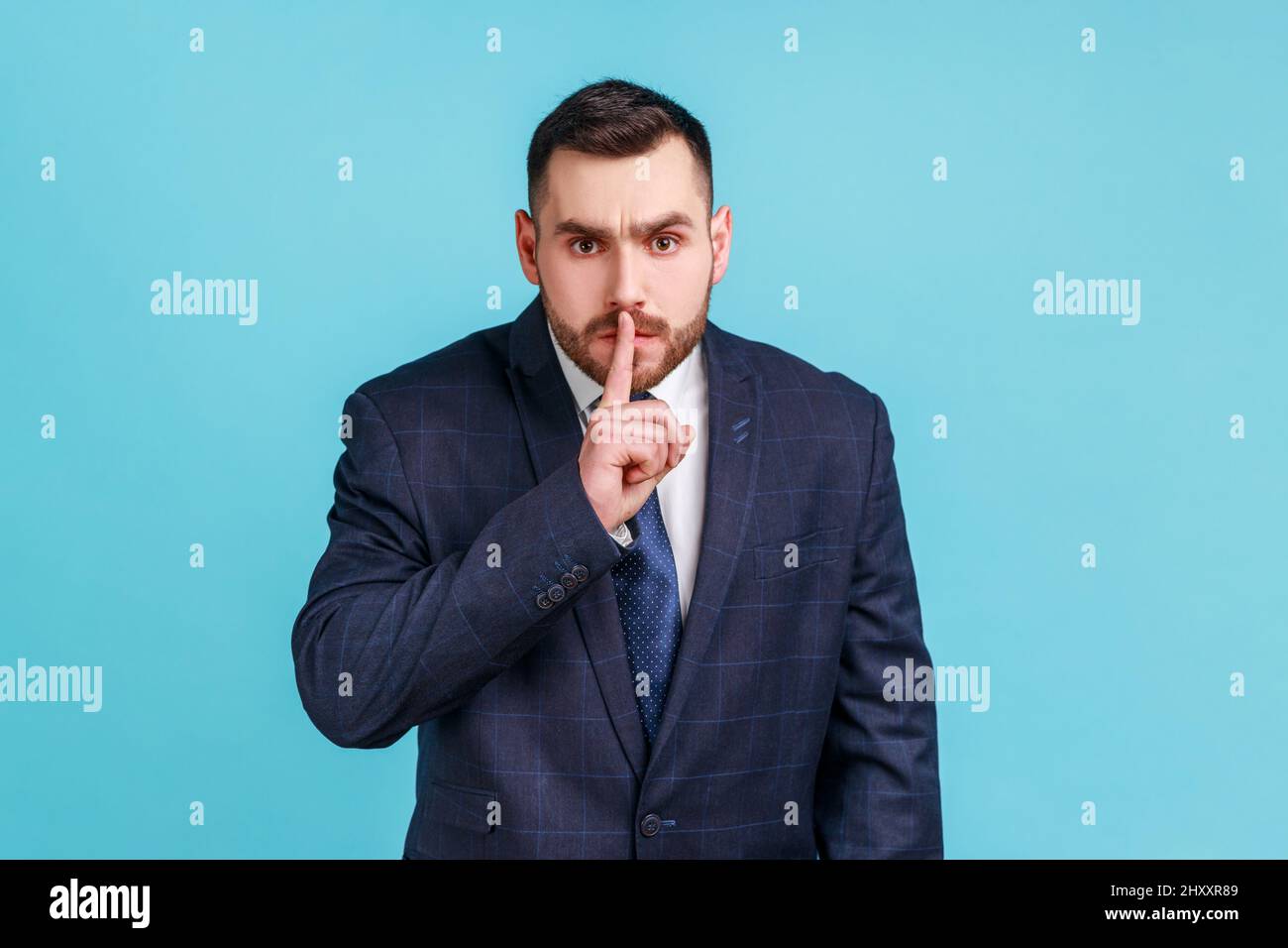 Portrait of bearded businessman wearing official style suit holding finger near lips and showing silence sign, keeping secrets, deceit. Indoor studio shot isolated on blue background. Stock Photo