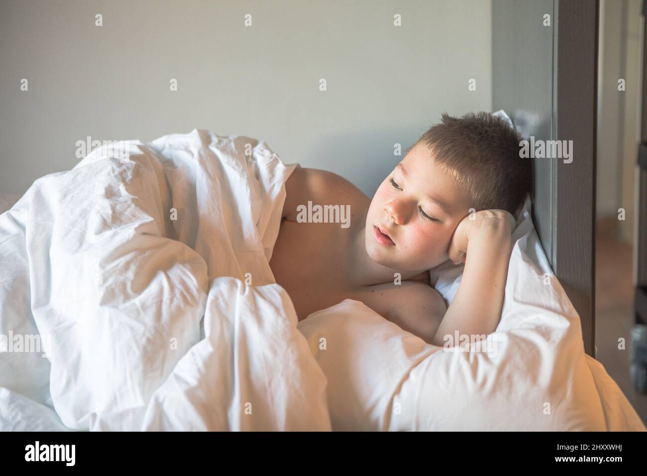 Kid 7 year old lying on bed, Sleepy child waking up the morning in his bed room with morning light, sad boy lies on a bed in the room Stock Photo