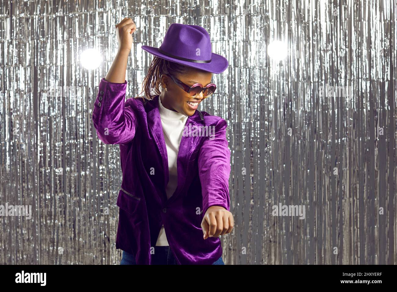 Cheerful African American woman in a purple hat, jacket and disco glasses dancing at a party Stock Photo