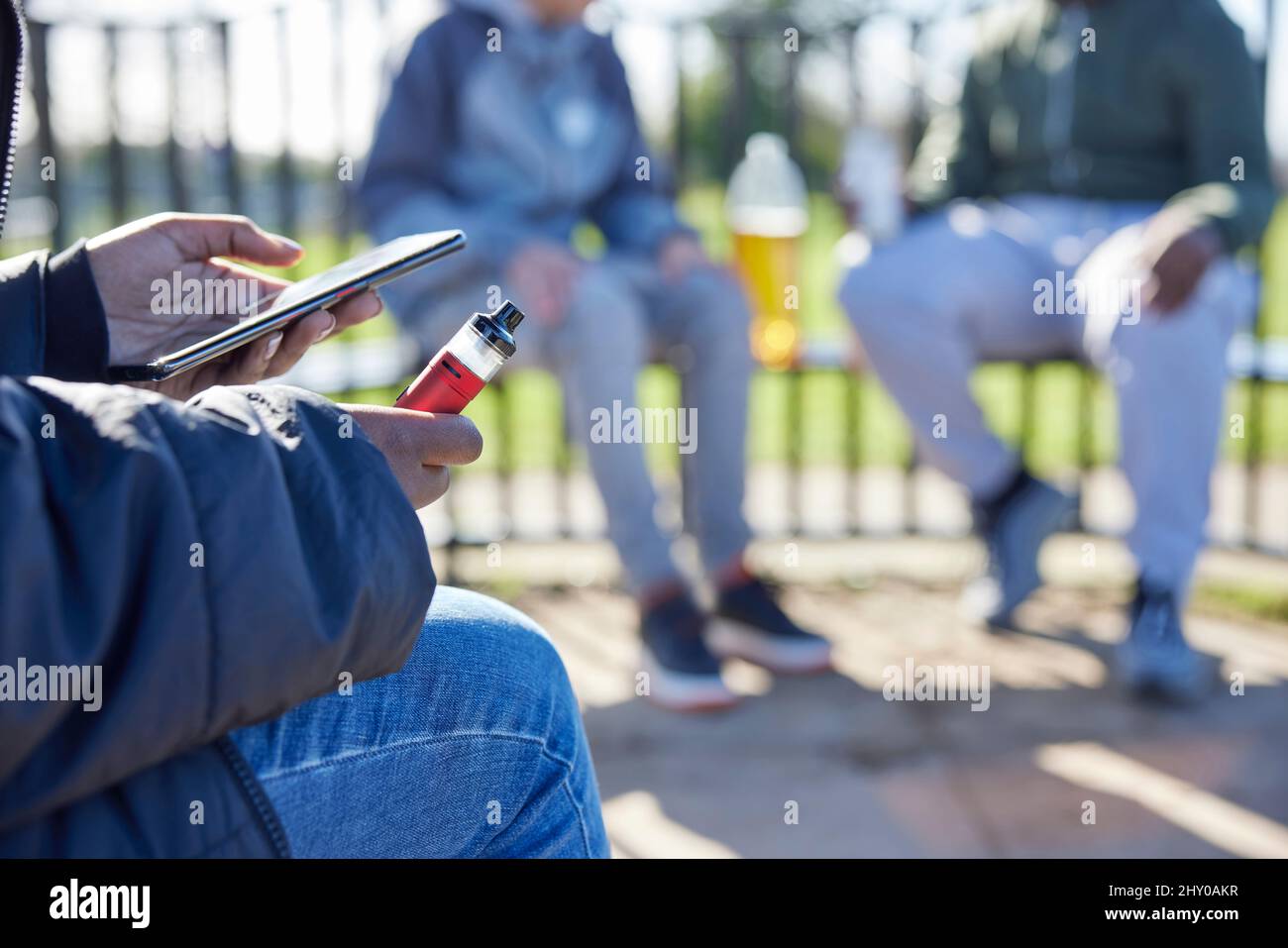 Close Up Of Teenagers With Mobile Phone Vaping and Drinking Alcohol In Park Stock Photo