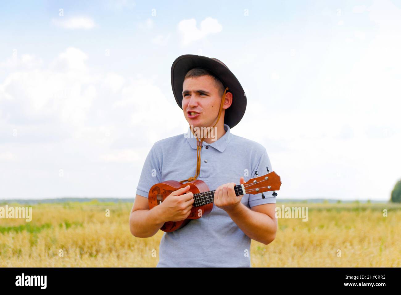 Summer fun. Young beautiful caucasian man playing ukulele outside. Handsome  hipster guy plays ukulele guitar on nature background. Male in a cowboy ha  Stock Photo - Alamy