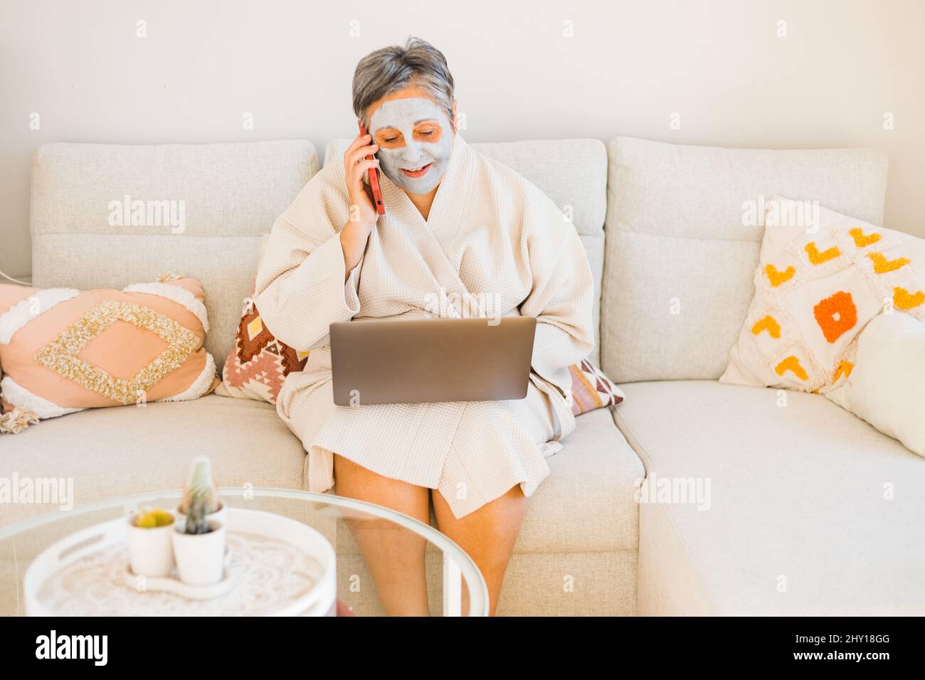 Woman in clay mask on face browsing netbook while sitting on comfortable couch and talking phone in light living room during skincare routine Stock Photo