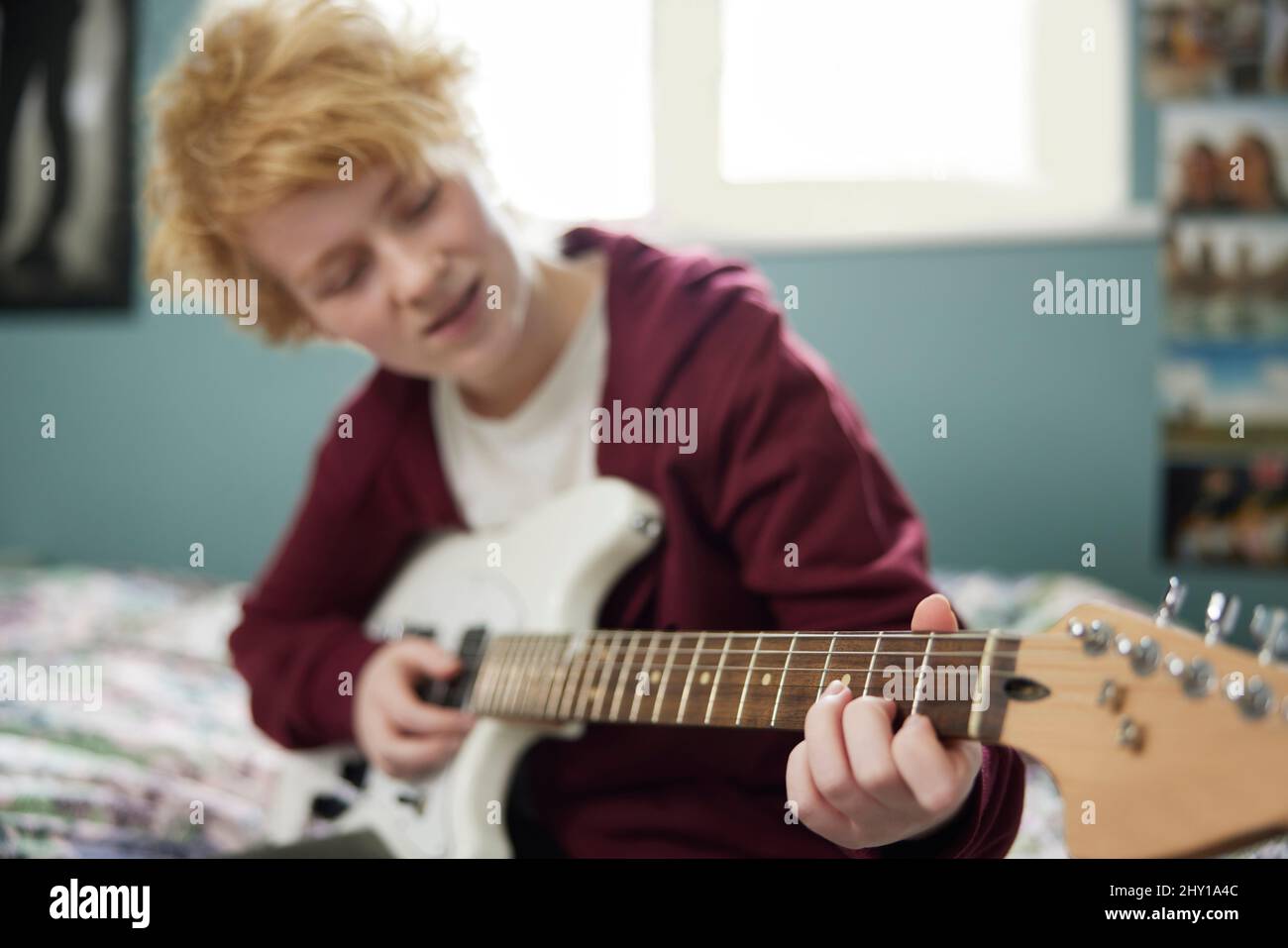 Teenage Girl Sitting On Bed Learning To Play Electric Guitar In Bedroom Stock Photo