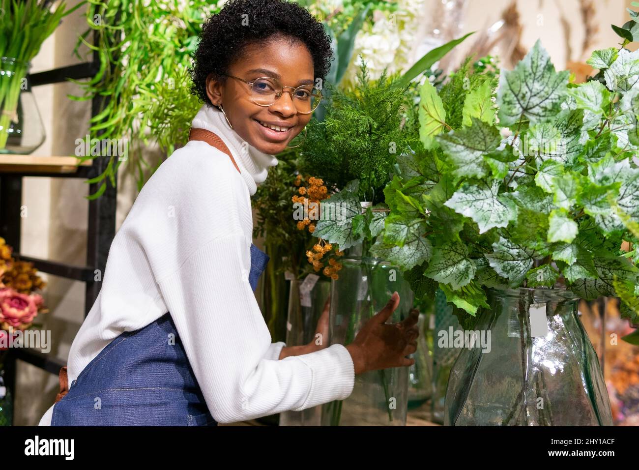 Positive African American female florist looking at camera while standing near green Tolmiea plants during work in modern floristry shop Stock Photo