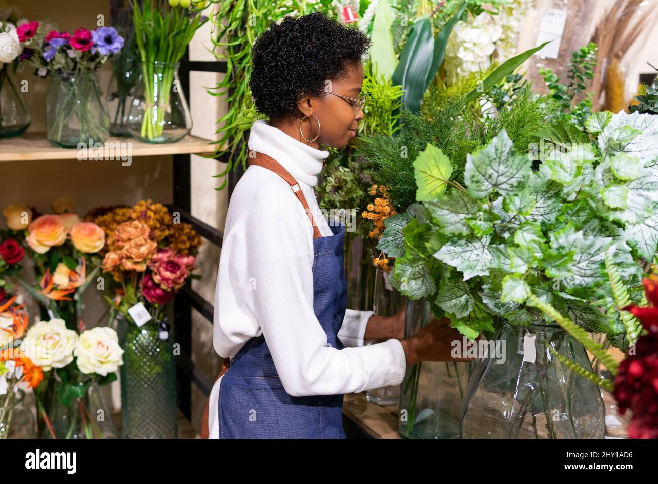 Positive African American female florist with eyes closed standing near green Tolmiea plants during work in modern floristry shop Stock Photo