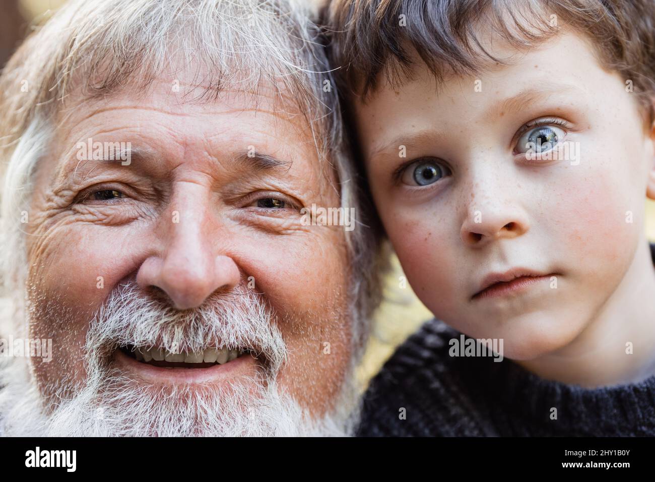 Positive aged man with gray beard and hair and cute smiling kid with blue eyes looking at camera in forest against blurred background Stock Photo