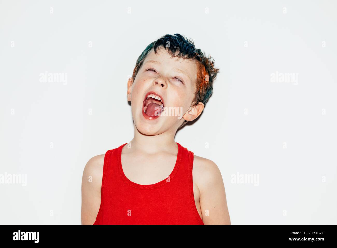 Mischievous boy with colorful dyed hair rolling eyes and showing tongue on white background in light room Stock Photo