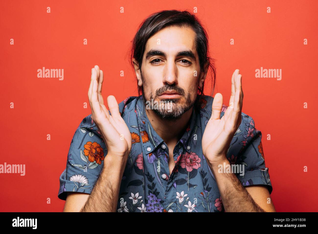 Bewildered man with beard in shirt looking at camera with raised hands while standing on red background in light studio Stock Photo