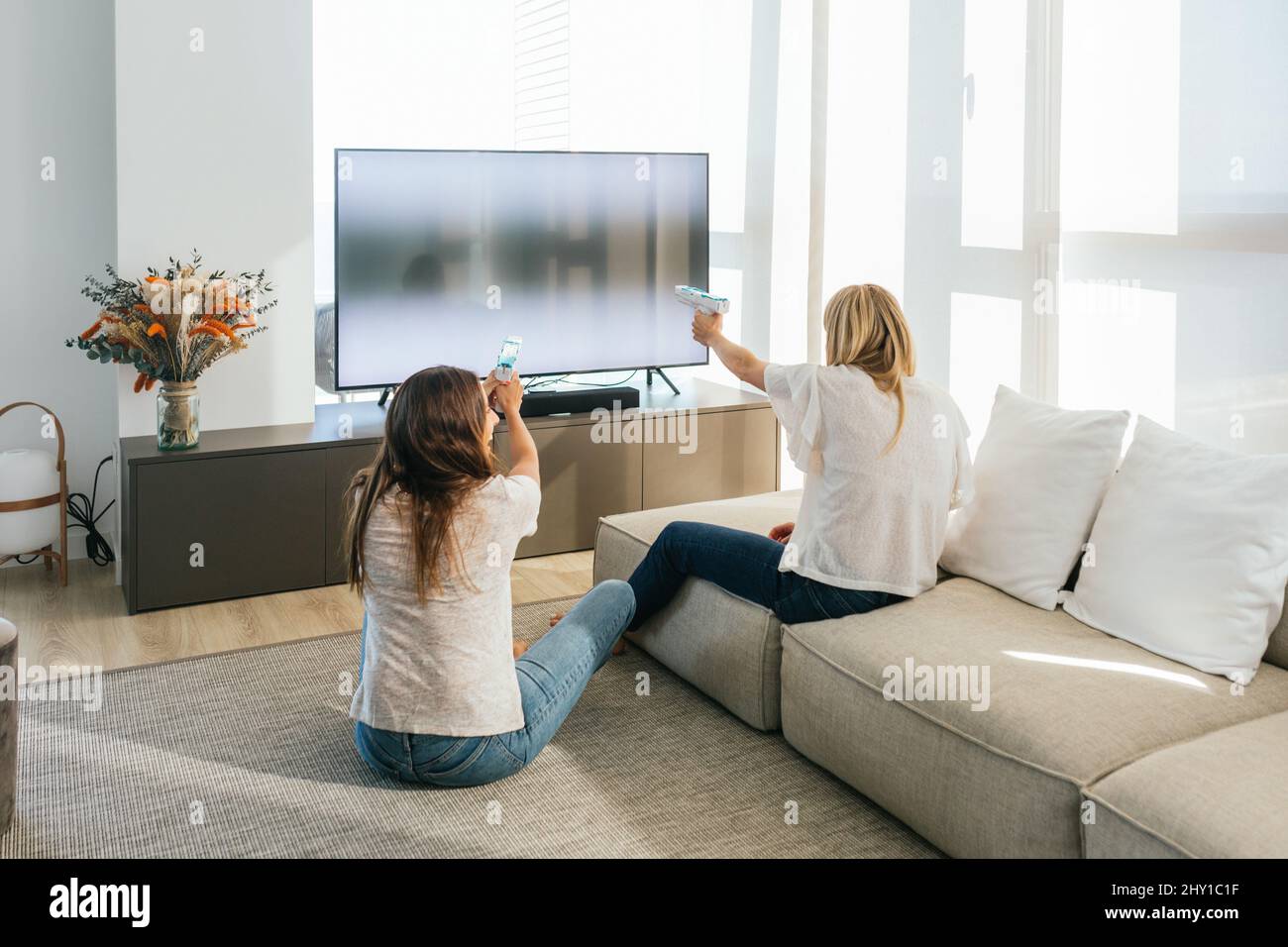 Back view of unrecognizable female friends with light pistols playing videogame on modern TV in light living room at home Stock Photo