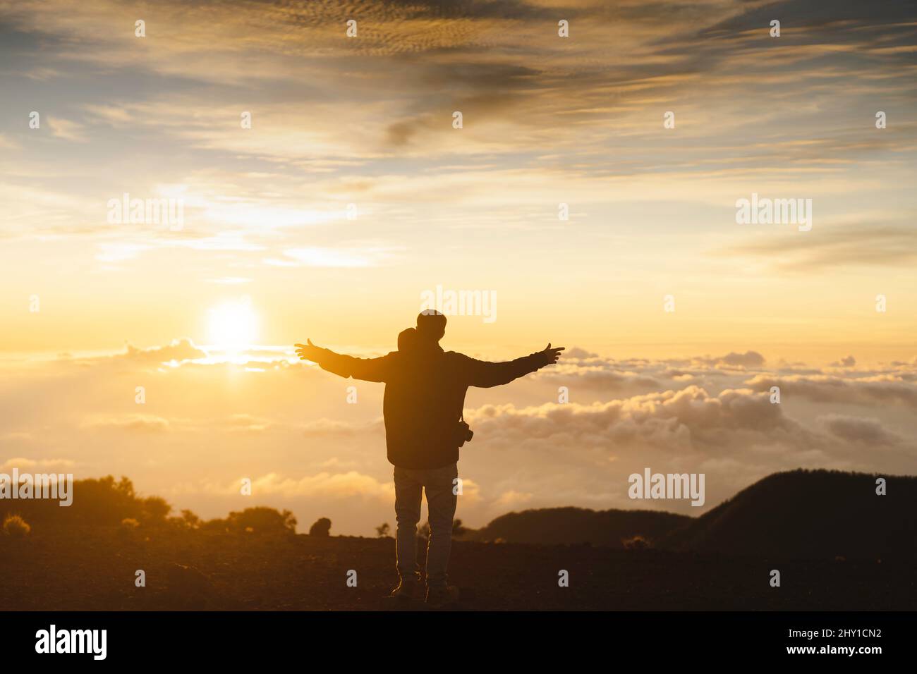 Full body back view of unrecognizable male traveler with spread arms while standing on top of mountain above cloudy sky at sundown time Stock Photo