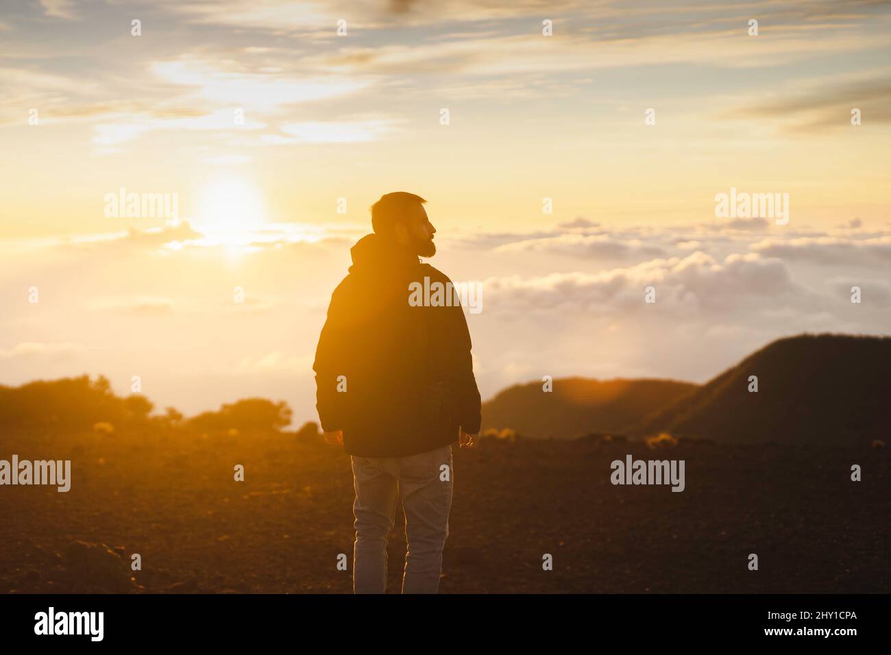 Back view of male traveler standing on top of mountain above cloudy sky at sundown time Stock Photo