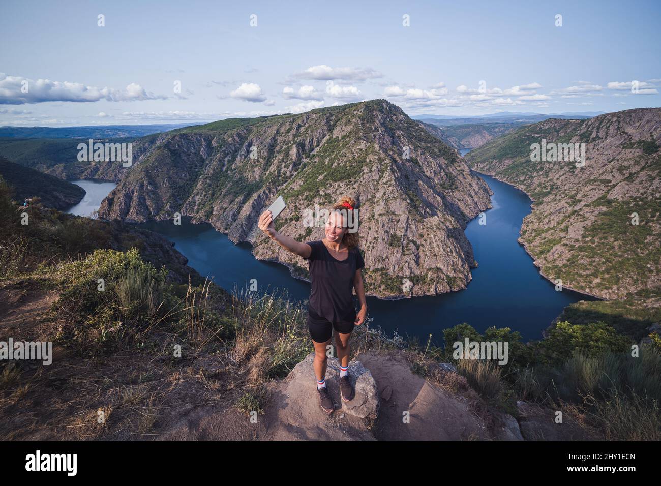 From above of full lenght adult female tourist in sportswear standing on top and taking selfie on smartphone while enjoying picturesque landscape Stock Photo