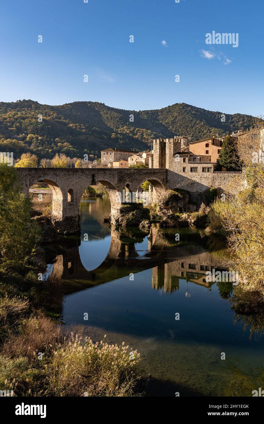 Aged stone bridge over river near ancient town on background of green forested mountains on sunny day in Spain Stock Photo
