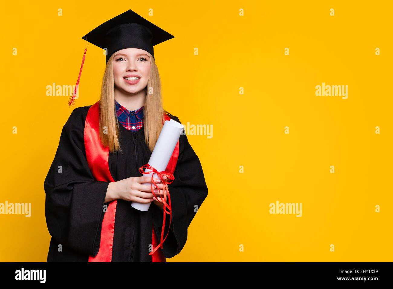 Portrait girl graduate with graduation hat and diploma on yellow background. Blonde young woman wearing graduation cap and ceremony robe holding Certificate tied with red ribbon. Education Concept Stock Photo