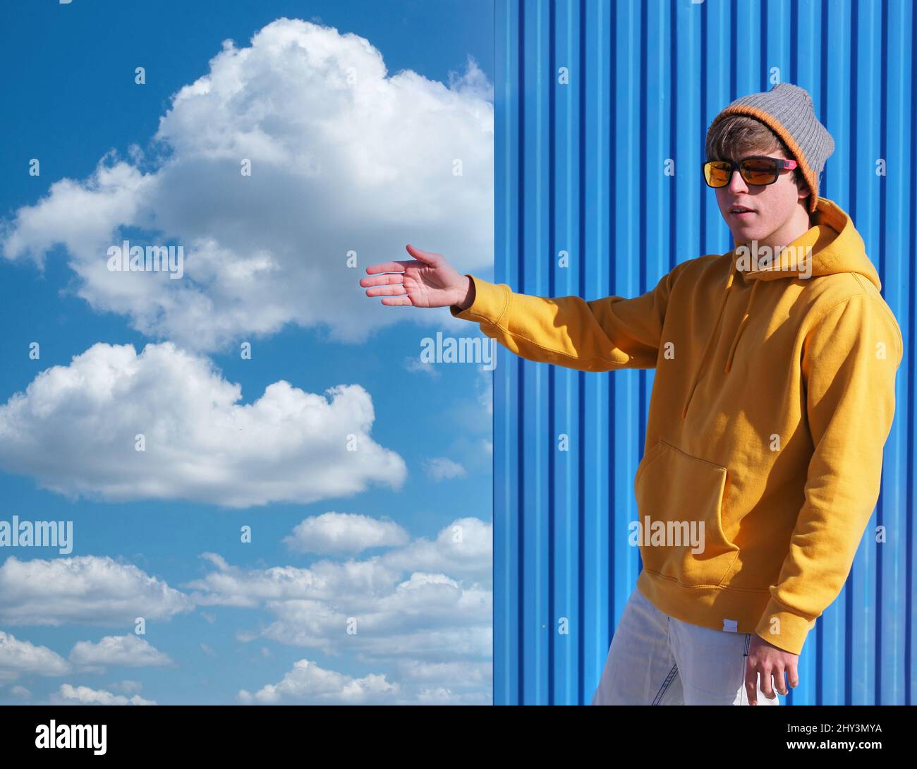 Cheerful young man in a yellow sweatshirt and wool hat pointing to the blue sky with clouds Stock Photo