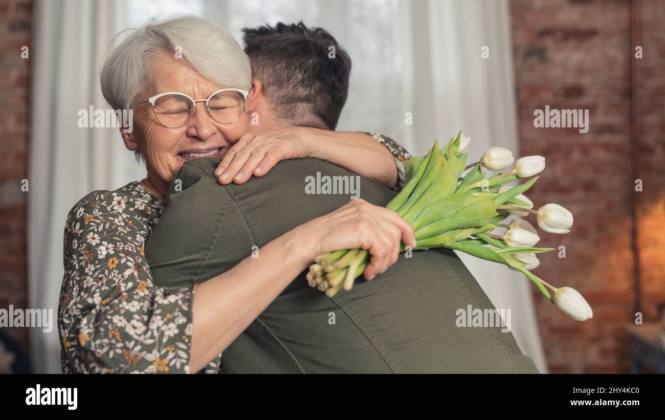 extremely happy caucasian grandmother hugs her grandson and holds white tulips bouquet on grandparent's day. High quality photo Stock Photo