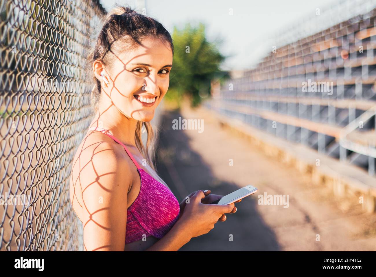Smiling caucasian young active woman looking at the camera and setting a music playlist on her smartphone Stock Photo