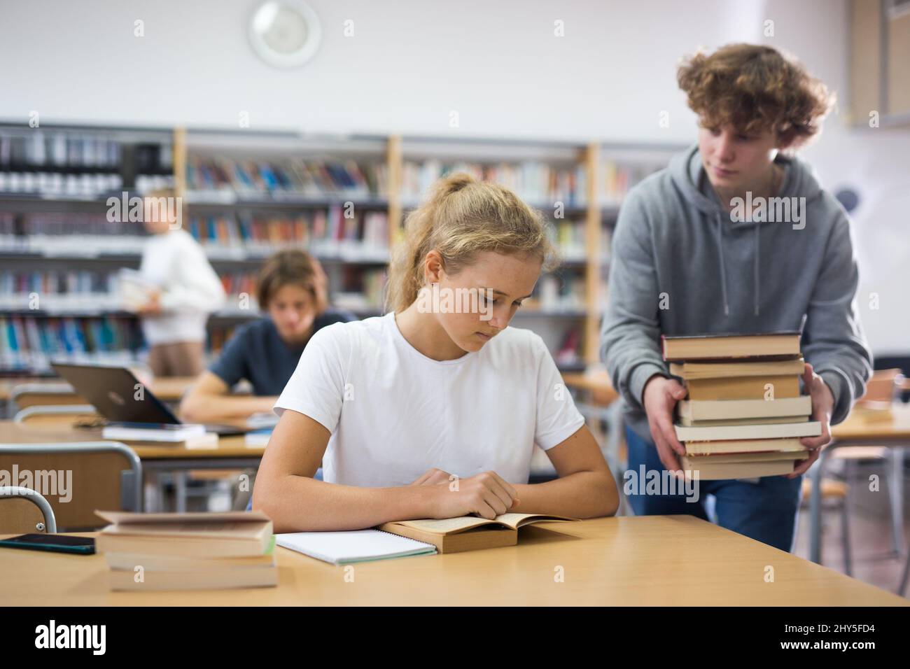 Teen brings stack of book to his girlfriend in school library Stock Photo