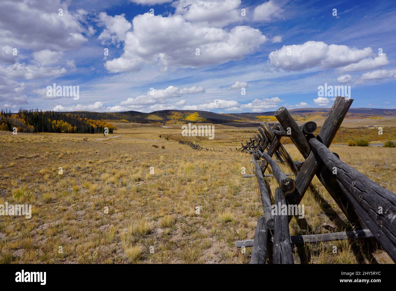 A wooden fence running along a meadow with mountains in the background covered in fall color. Stock Photo
