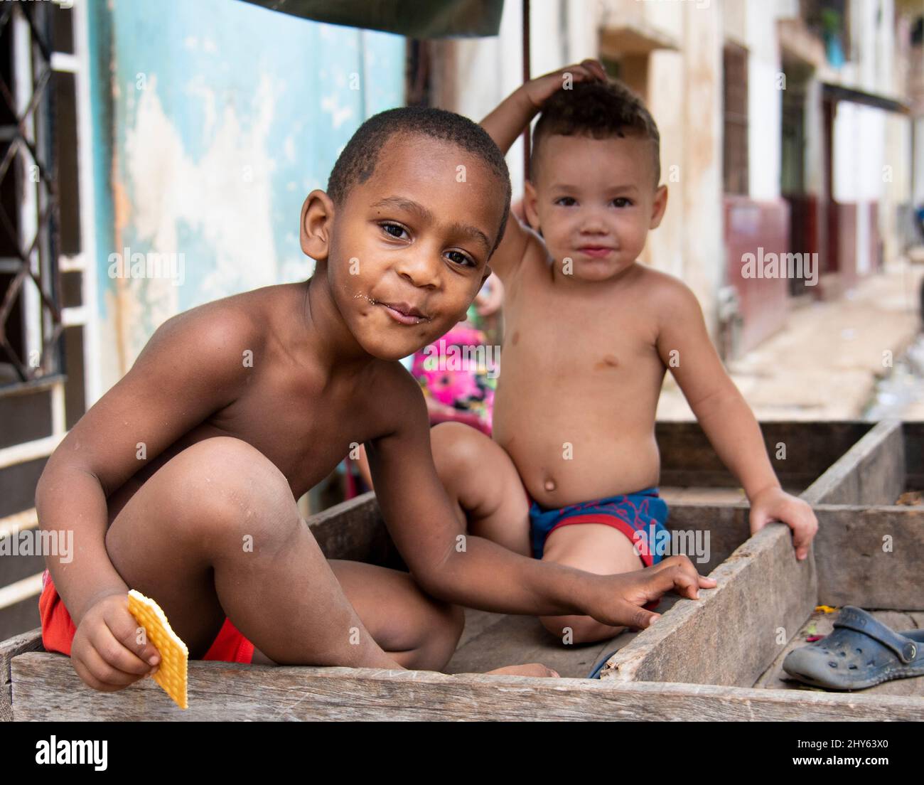 Young cuban boy eats a cookie while his friend looks and smiles at the camera while sitting on a cart in a neighborhood in Havana, Cuba. Stock Photo
