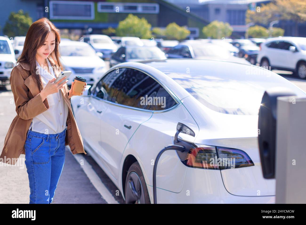 Smiling young woman standing on city parking near electric car, charging automobile battery from small city station, drinking coffee and using smartph Stock Photo
