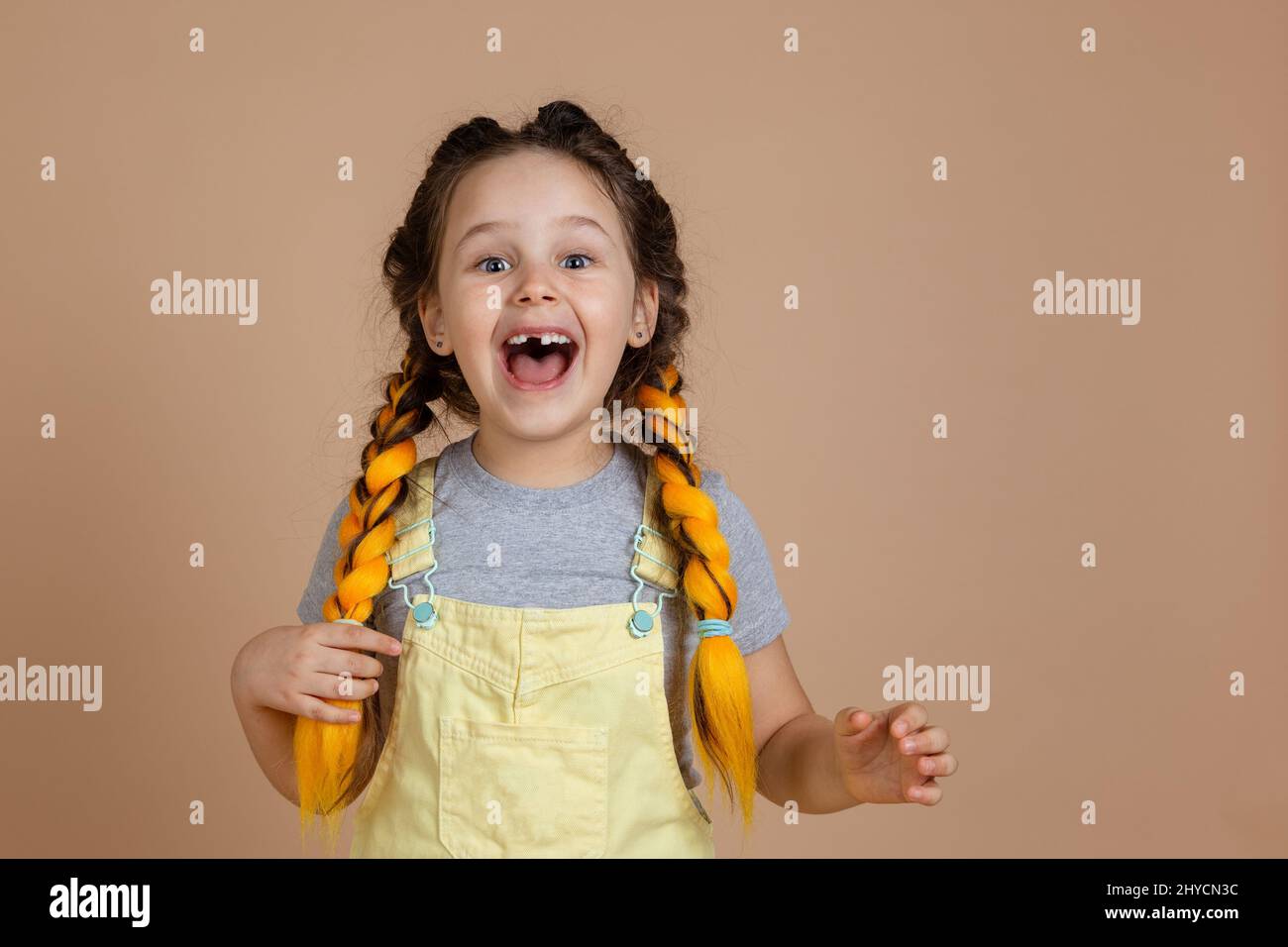 Fascinated young nice female holding yellow kanekalon pigtail with hand, smiling with missing tooth looking at camera wearing yellow jumpsuit and gray Stock Photo