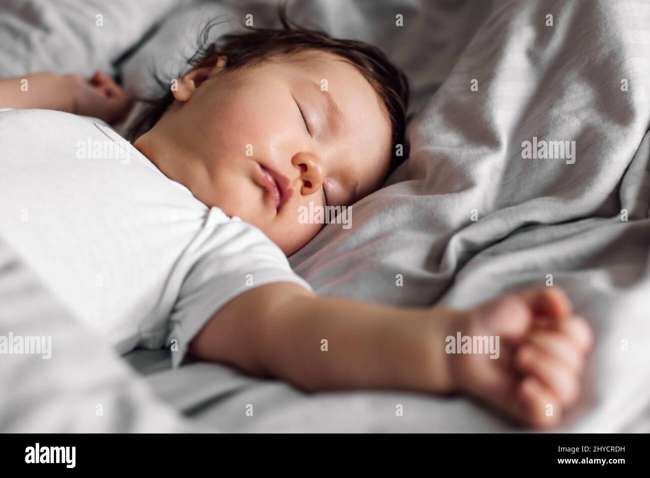 Portrait of baby in sleepwear lying under blanket on bed at home. Sleeping infant child in bedroom, see sweet dream. Grey background, soft focus, free Stock Photo
