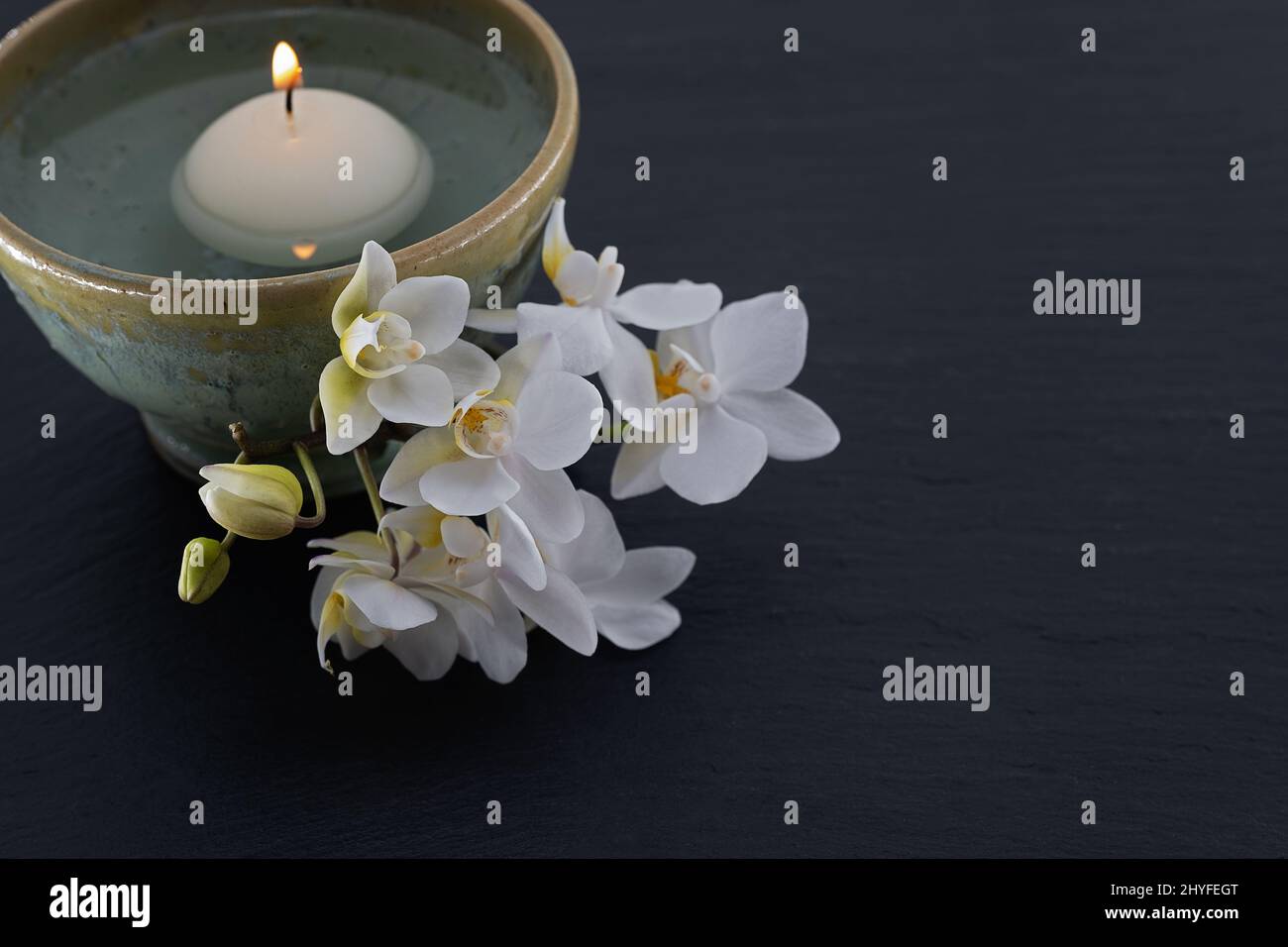 white floating candle in handmade ceramic bowl with a sprig of orchids on black slate board Stock Photo