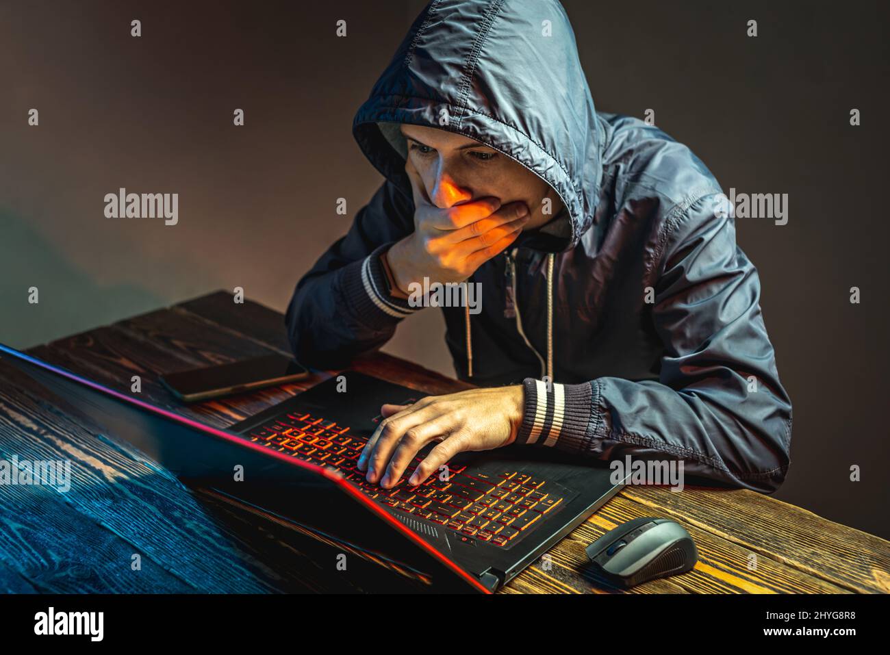 A hacker in a hood with a phone is typing on a laptop keyboard in a ...