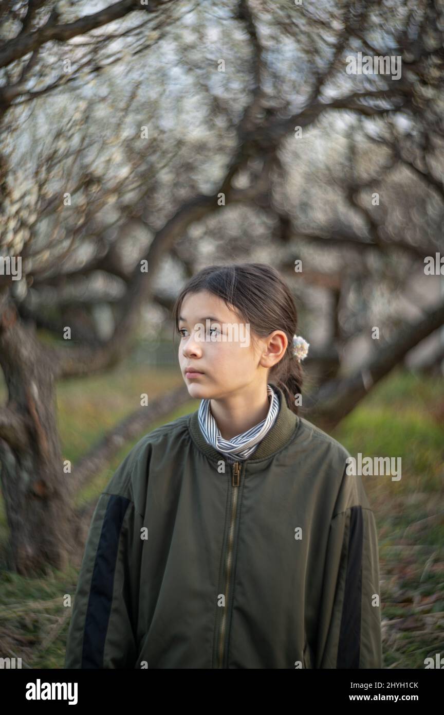 Profile portrait of a happy pre teen girl in a park. Child wearing a green jacket and turtleneck. Blooming trees in the background. Springtime. Stock Photo