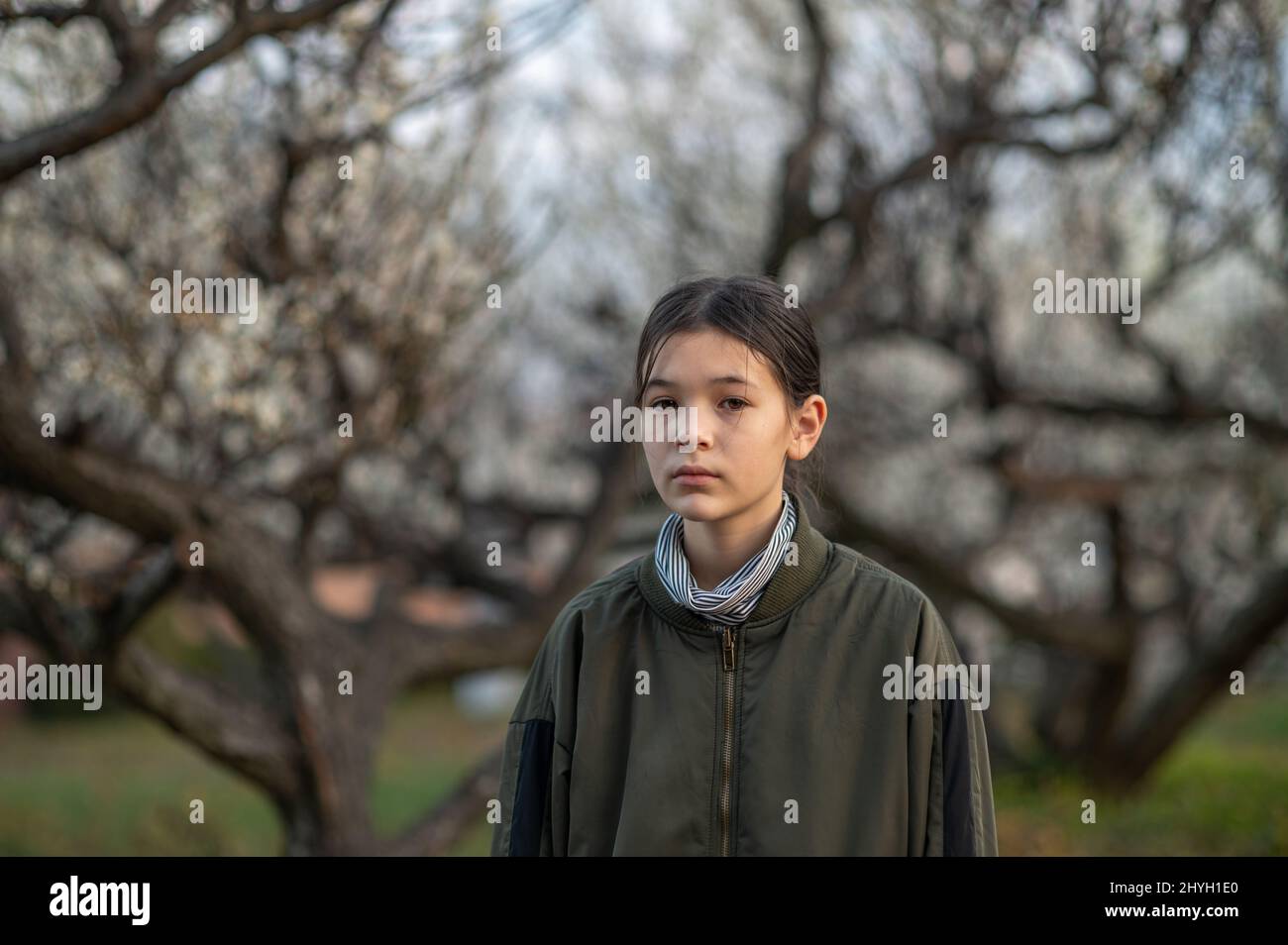 Portrait of a sad and sick pre teen girl in a park. Child wearing a green jacket and turtleneck. Blooming trees in the background. Springtime. Stock Photo