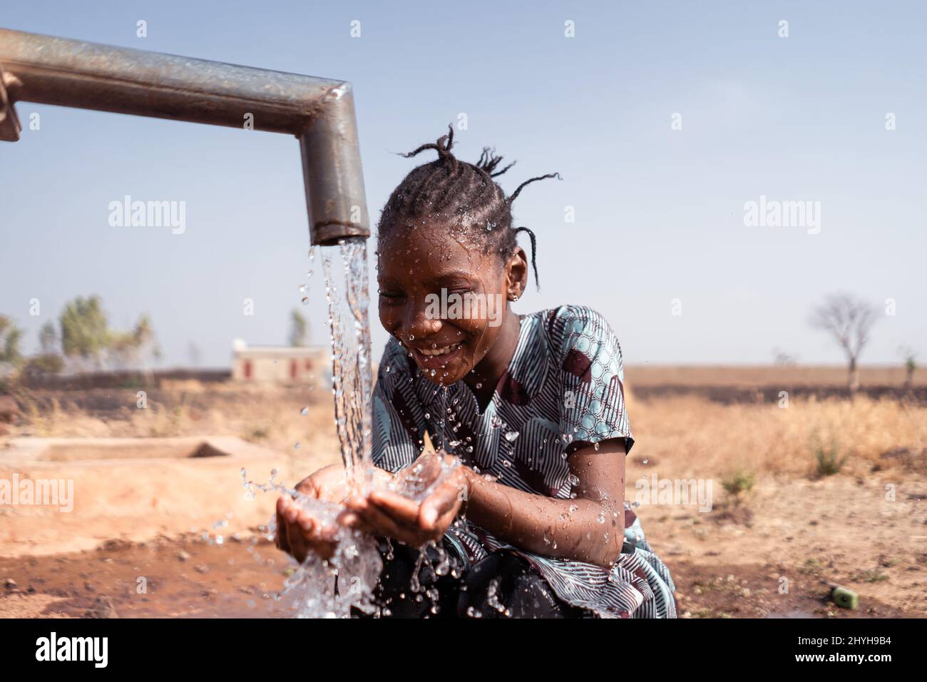 Happy little African girl crouching in front of a waterhole in an arid environment somewhere in the subsaharn region, drinking lots of fresh water fro Stock Photo