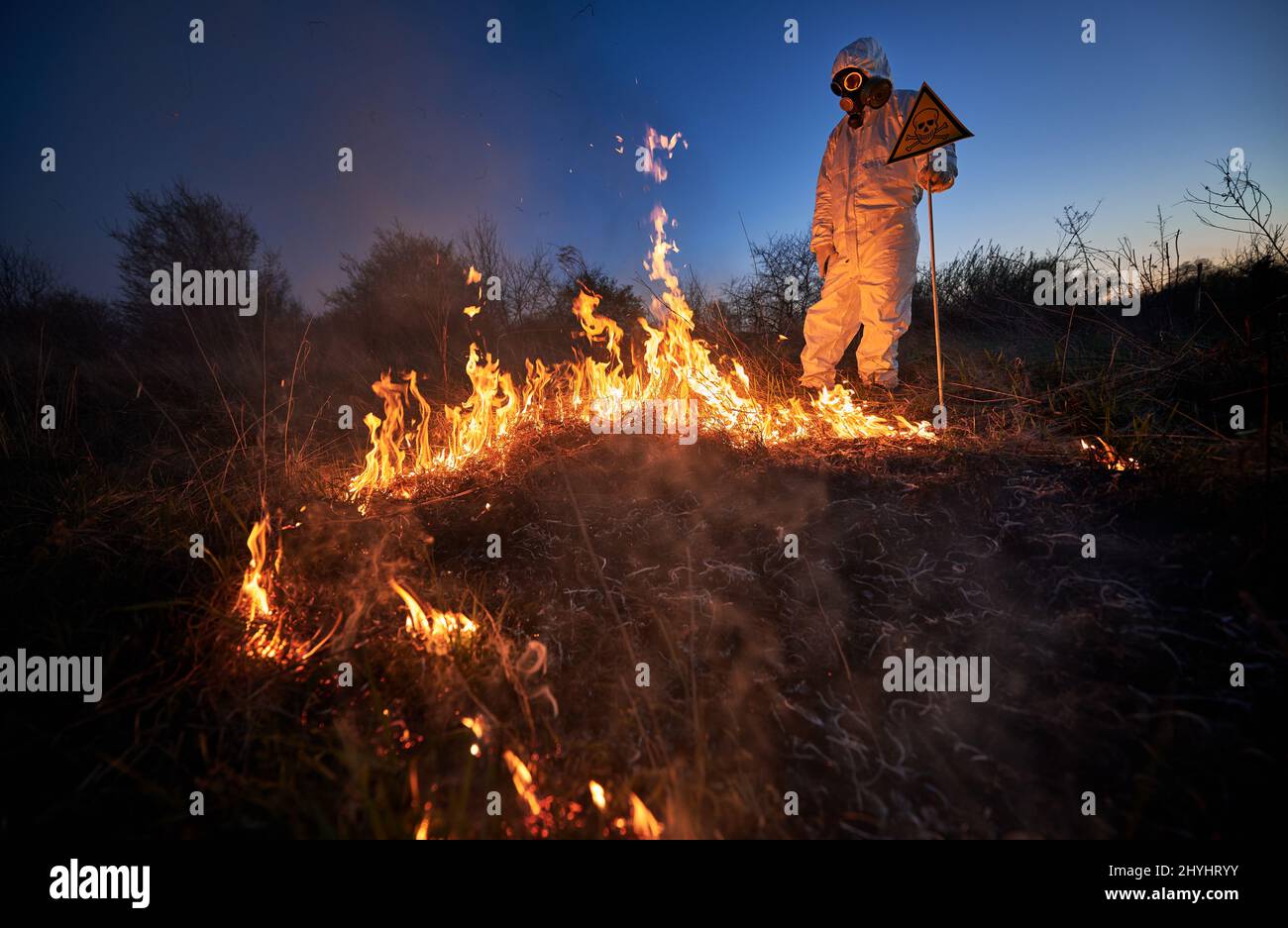 Firefighter ecologist extinguishing fire in field at night. Man in protective suit and gas mask near burning grass with smoke, holding warning sign with skull and crossbones. Natural disaster concept. Stock Photo