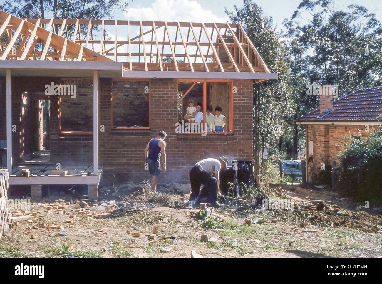 Sydney Australia 1959: A young family and friend look on while builders work on the new backyard of a red brick home in Turramurra on Sydney's north shore in Australia. In the days before lightweight excavators, large tree stumps were removed by being set on fire and slowly burnt out. Grace Bros (department store) Home Plans Service, established in association with the Sunday Telegraph Newspaper, Sydney, was launched in April 1954 and this home is based on one of those plans. See also Alamy Image ID 2BWEYMX for the completed home Stock Photo