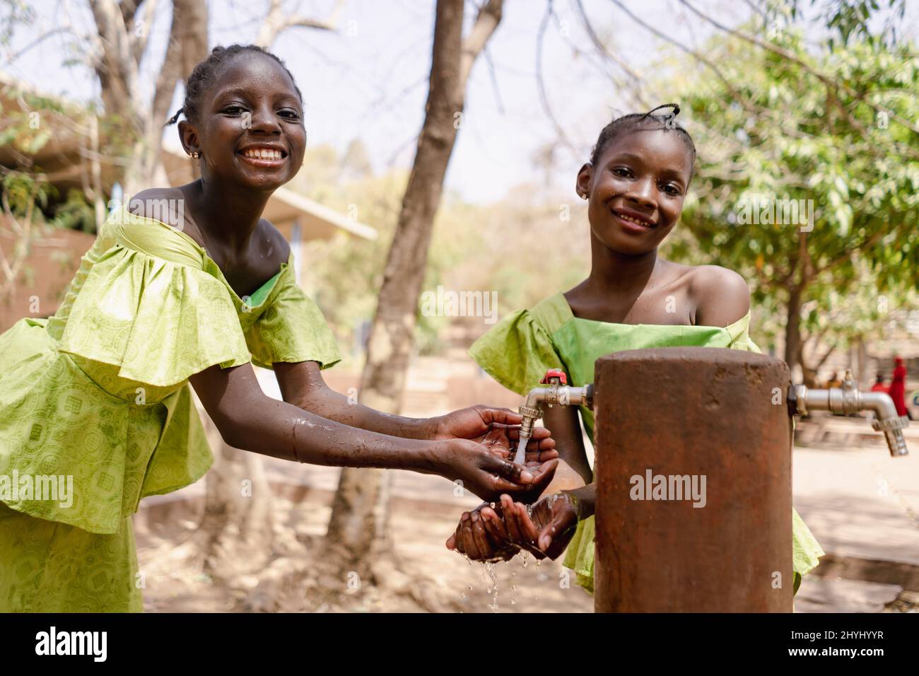 Two charming African girls in beautiful traditional dresses refresh themselves with water from a public village tap; human right to water and sanitati Stock Photo