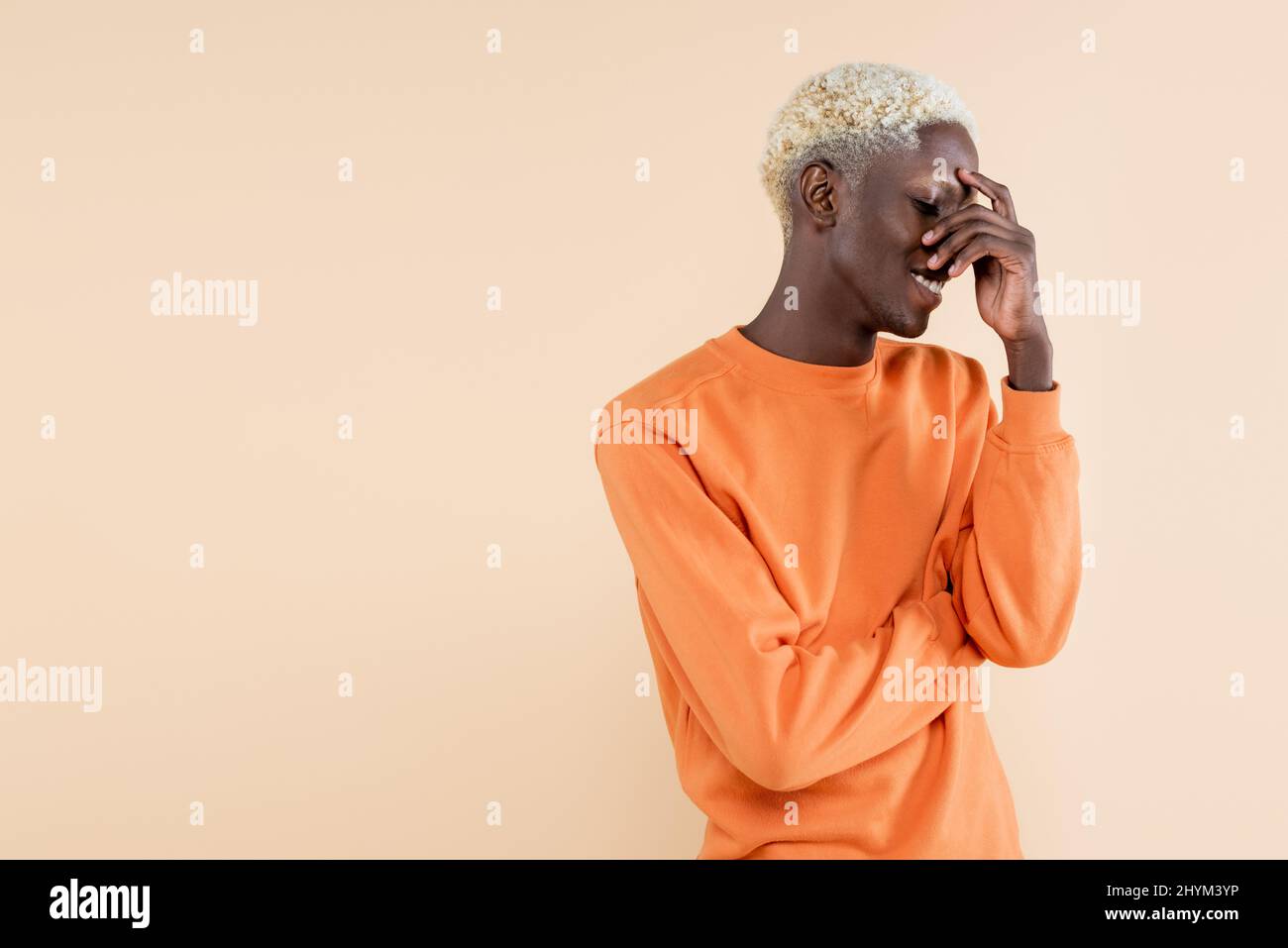 shy african american man in orange sweatshirt smiling and covering eyes isolated on beige Stock Photo