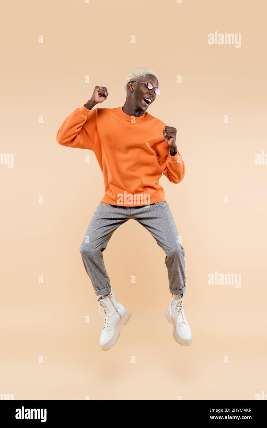 full length of excited african american man in orange sweatshirt and sunglasses jumping on beige Stock Photo