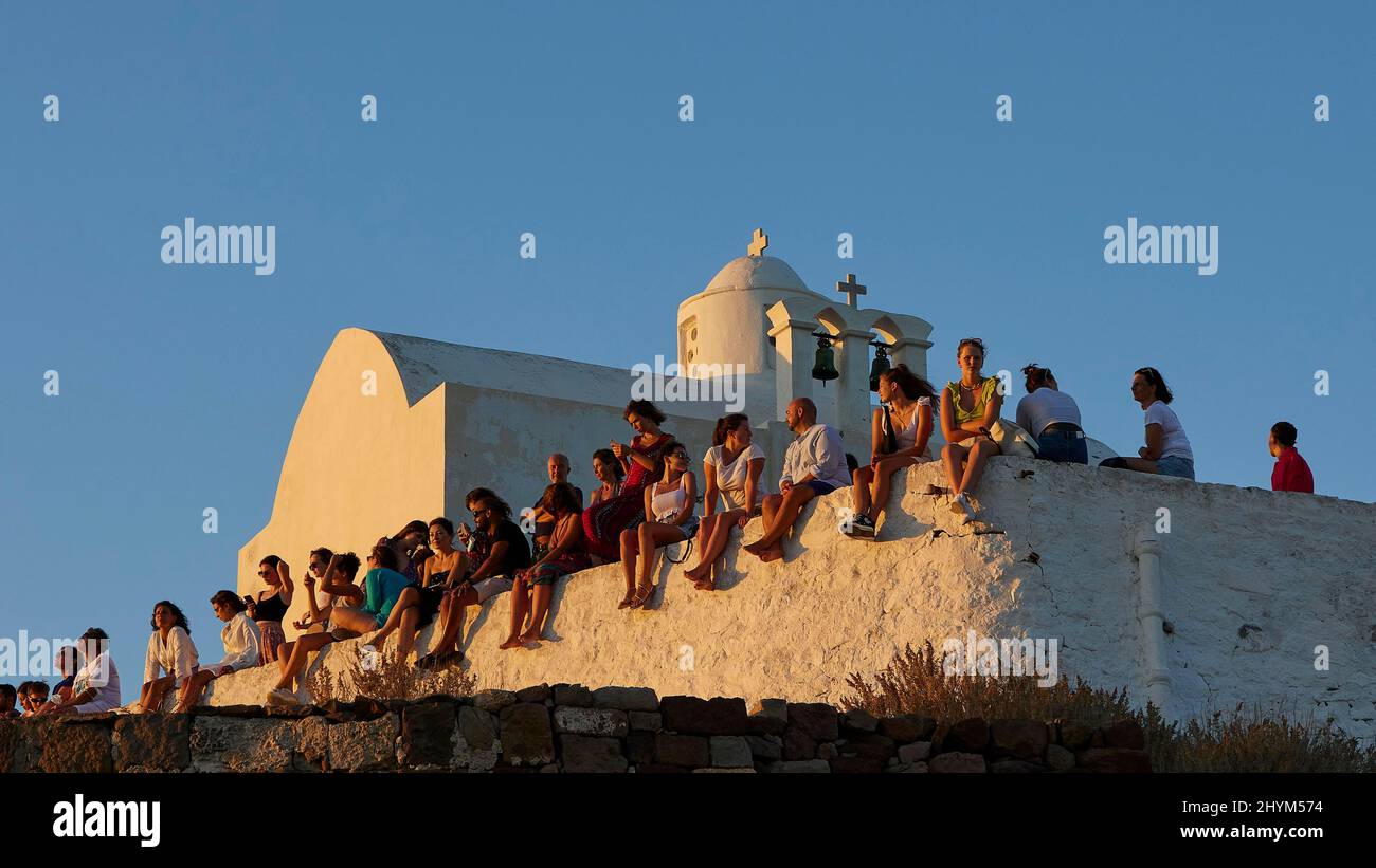 Evening atmosphere, group of people sitting on a wall, small white chapel, Venetian fort, Plaka, Milos Island, Cyclades, Greece Stock Photo