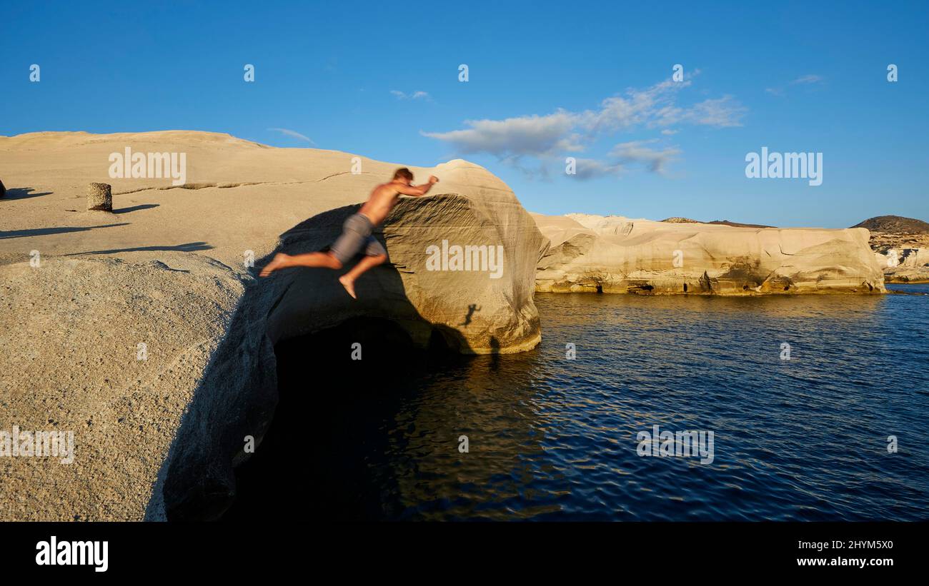 Evening light, man jumps from rocks into water, bizarre tuff rock, blue sky, grey-white cloud, sea dark blue, Sarakiniko Bay, Milos Island, Cyclades Stock Photo