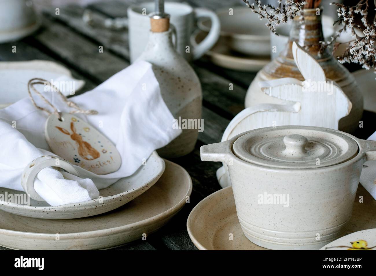 Rustic Easter table setting with empty craft ceramic tableware, cooking pot, plates and bowls, Easter bunny rabbit decorations, vase on wooden table. Stock Photo