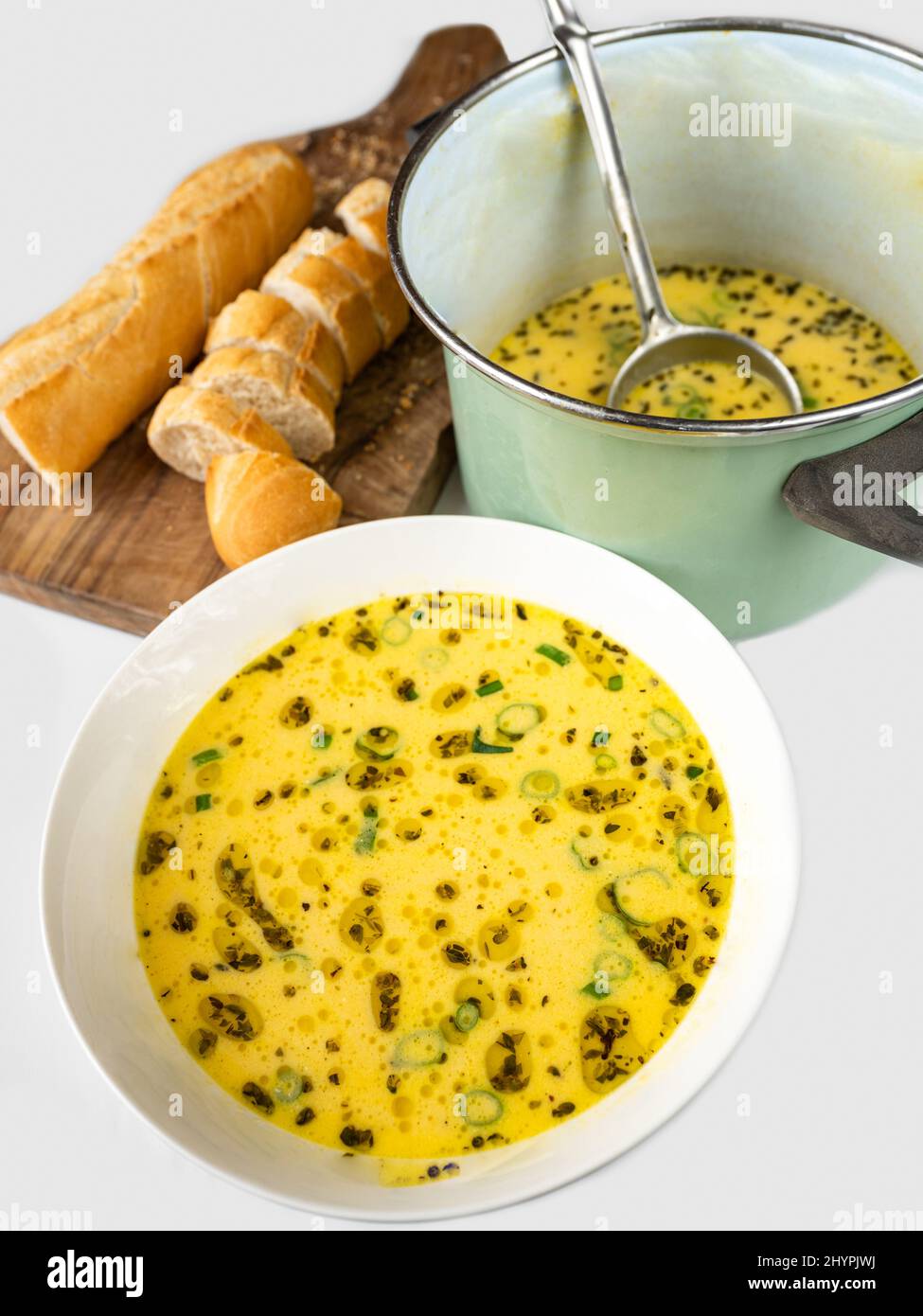 Cheese soup with oregano herb and green onion shoot in white plate and in green pot, sliced bread, all on white background. Stock Photo