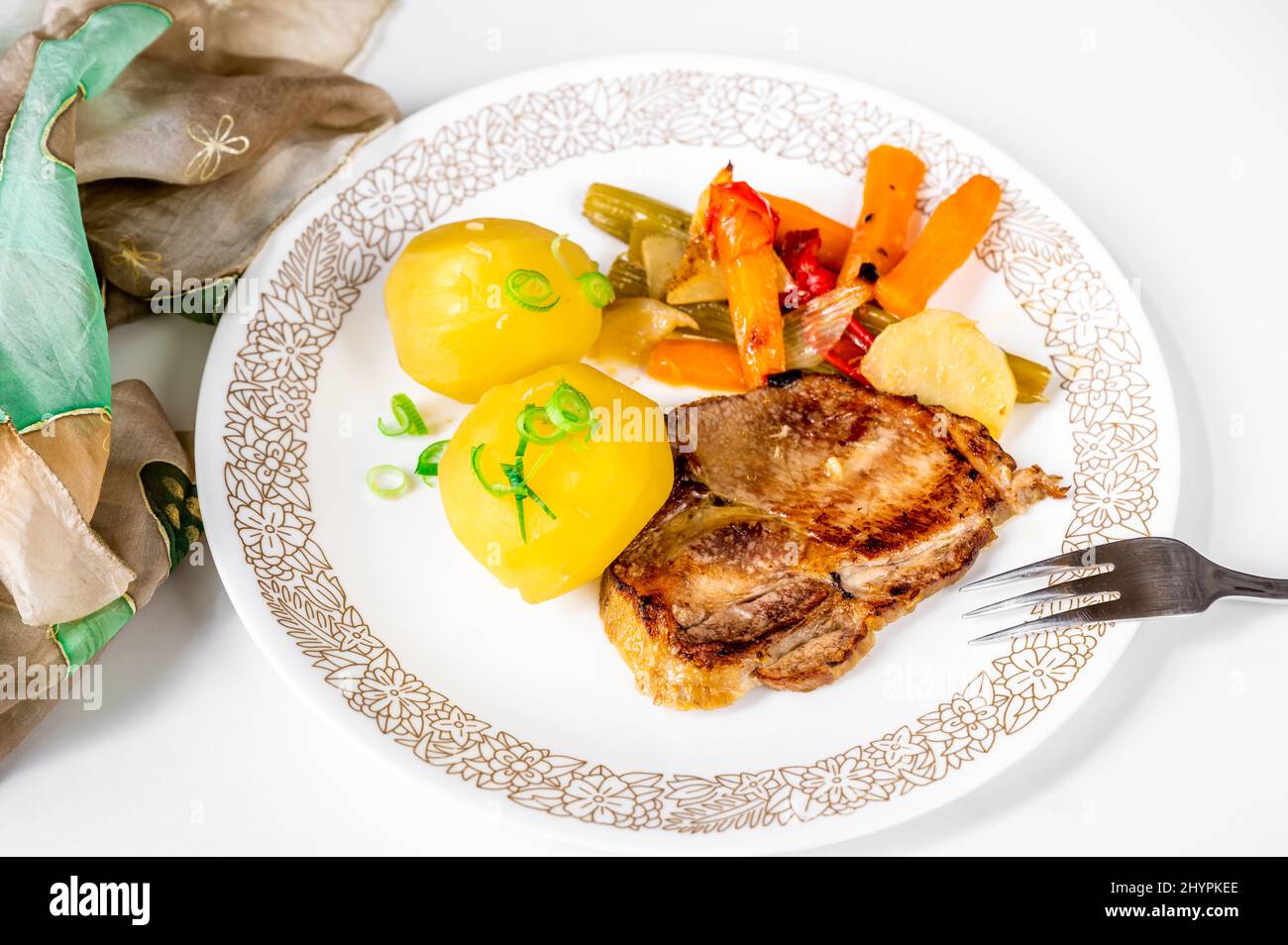 Roasted pork steak, boiled potato and baked vegetable on white plate with fork, on white background with towel, closeup. Stock Photo