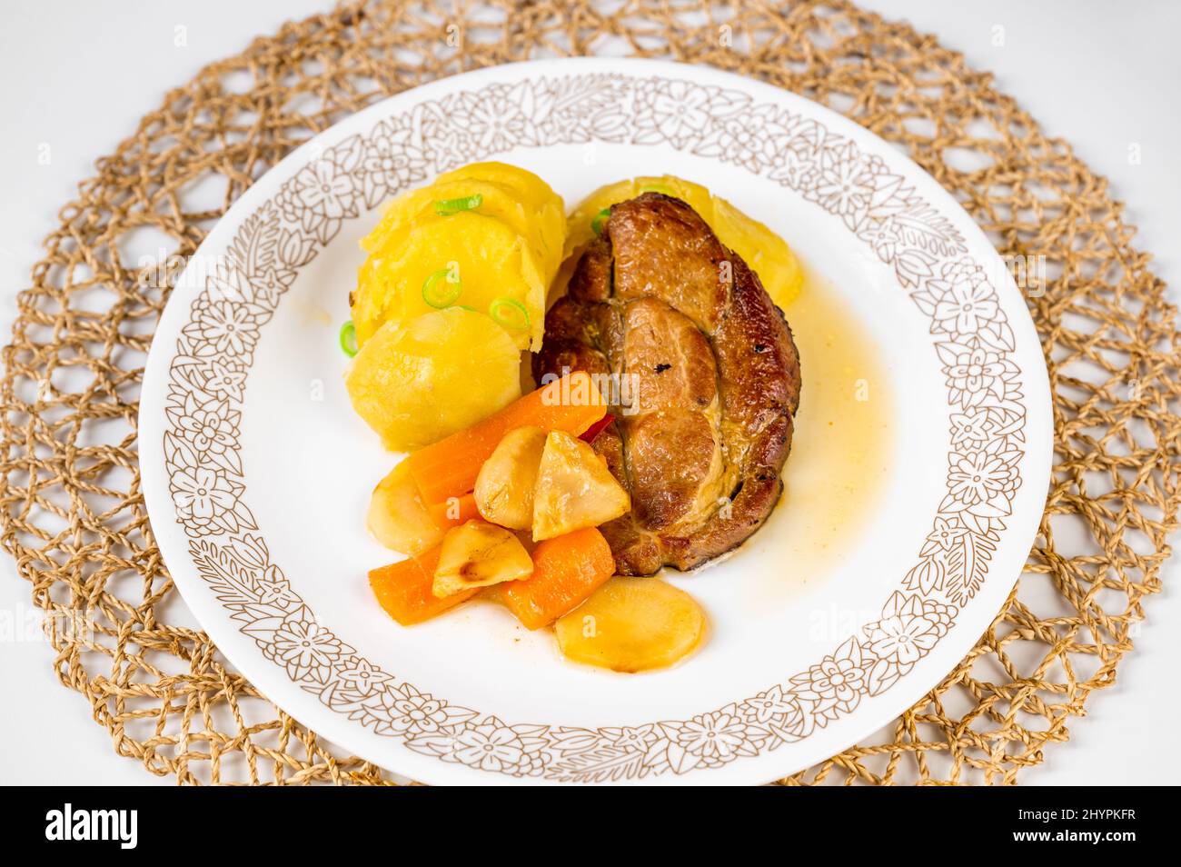 Portion of roasted pork neck steak, boiled potato, baked vegetable on white plate on bamboo pad on white background, closeup. Stock Photo