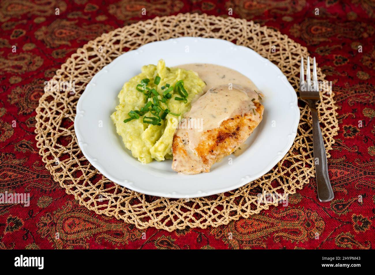 Chicken pocket with cheese and ham, fragrant rosemary sauce and mashed potato with mashed pea on white plate, bamboo pad, fork on red oriental tablecl Stock Photo