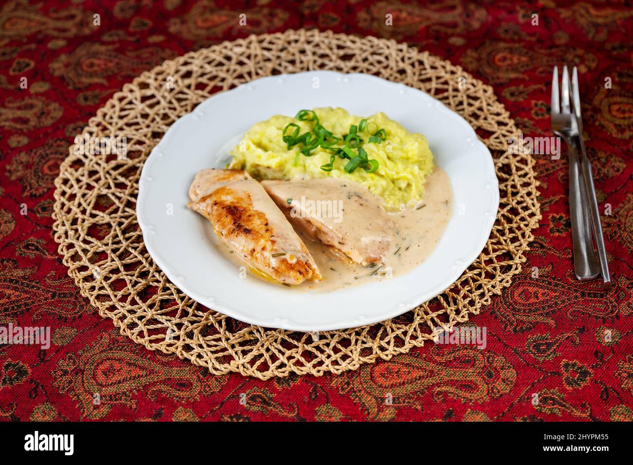 Sliced chicken pocket stuffed with cheese and ham, fragrant rosemary sauce, mashed potato with mashed pea on white plate, bamboo pad, fork on red orie Stock Photo