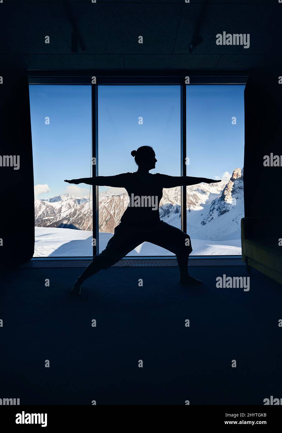 Young woman silhouette doing meditation yoga warrior virabhadrasana pose with in the room with panoramic view of a beautiful mountain landscape. Healt Stock Photo