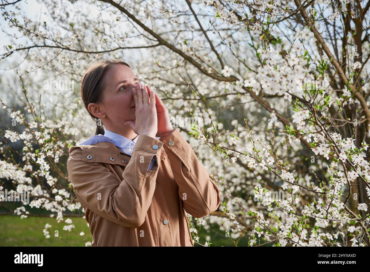 Woman allergic suffering from seasonal allergy at spring, posing in blossoming garden at springtime. Young woman sneezing in front of blooming tree. Spring allergy concept Stock Photo