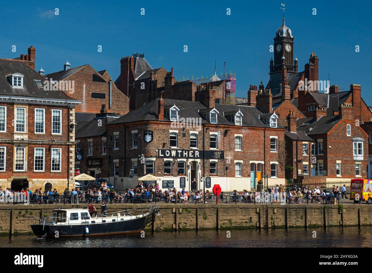 Customers drinking, eating alfresco in sun at busy riverside pub & leisure boat moored - River Ouse, King's Staith, York, North Yorkshire, England. Stock Photo