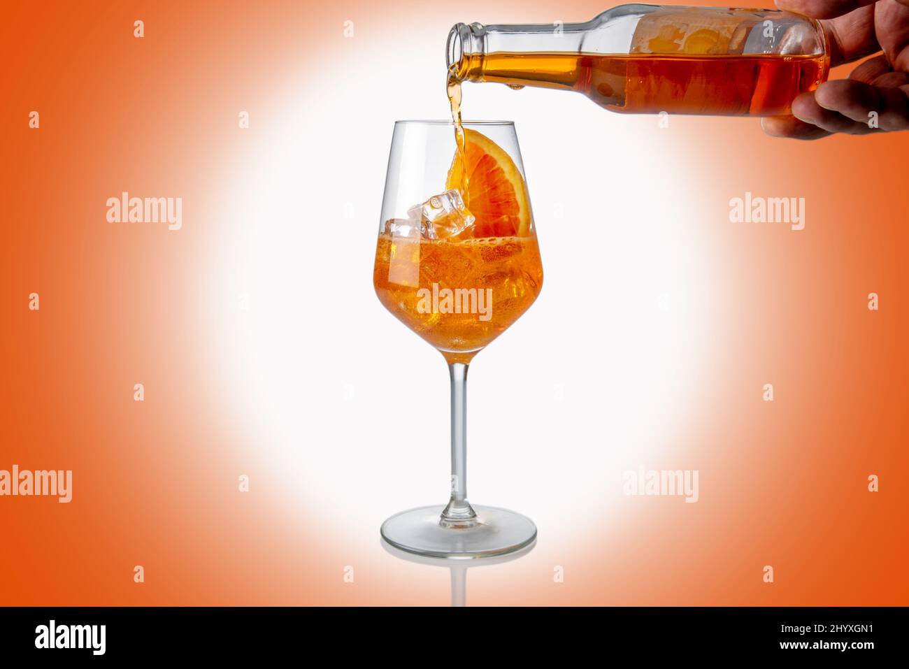 Alcoholic Aperol Spritz Cocktail poured from glass bottle into glass with ice cube and orange slice, spot white light on orange background Stock Photo